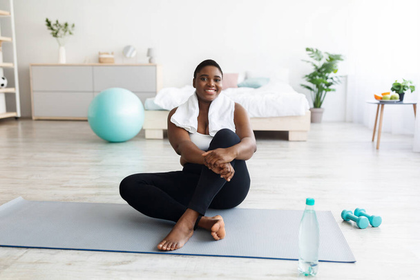 Smiling overweight young black woman sitting on yoga mat, resting after home fitness, copy space - 写真・画像