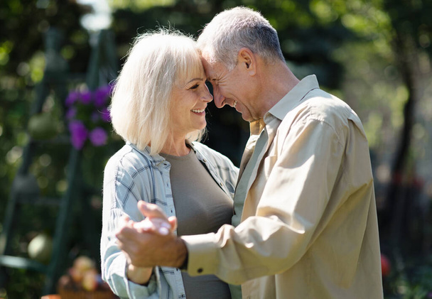 Zarte ältere Eheleute tanzen an einem Frühlingstag im Garten, berühren die Stirn und lächeln, ruhen zusammen im Freien - Foto, Bild