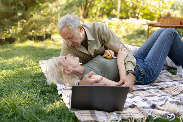 Tender married senior couple enjoying time together, spending day outdoors in their garden, having picnic on lawn - Foto, afbeelding