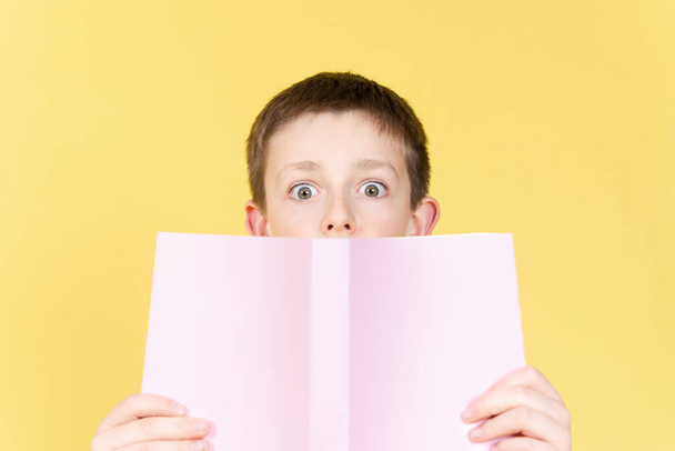 Verrast jongen achter een boek kijken recht naar camera met open ogen geïsoleerd op gele achtergrond - Foto, afbeelding