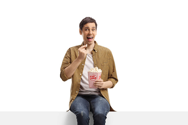 Excited young man sitting on a panel and eating popcorn isolated on white background - 写真・画像