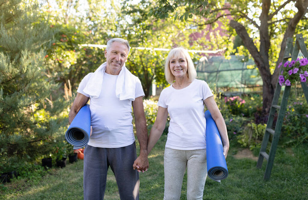 Concepto de estilo de vida saludable. Felices cónyuges mayores deportistas sosteniendo esteras de yoga y sonriendo a la cámara después de hacer ejercicio al aire libre - Foto, Imagen