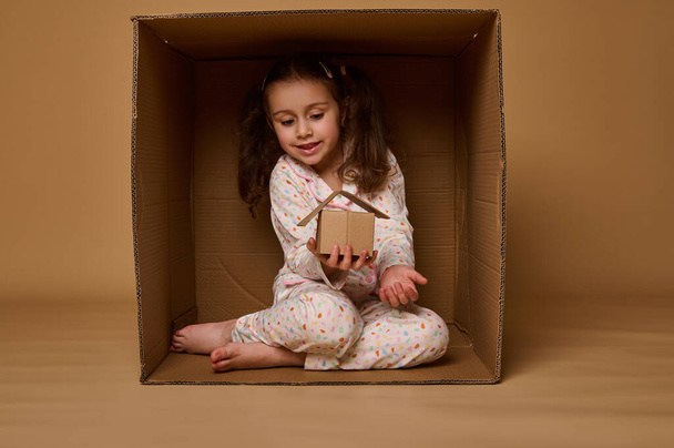 Adorable Caucasian little girl hiding inside a box and holding a craft cardboard house model. Real estate, housing, insurance and social secure concept with copy space for advertisement - Foto, Imagen