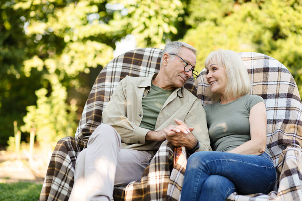 Любящие пожилые супруги отдыхают в своем саду в сельской местности, сидят в плетеных стульях и смотрят друг на друга - Фото, изображение