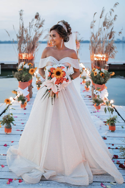 A beautiful Asian bride in a wedding dress poses at sunset. A bouquet of flowers and a long veil. Wedding decor in traditional bright colors. Filipino customs and holidays. - Fotografie, Obrázek