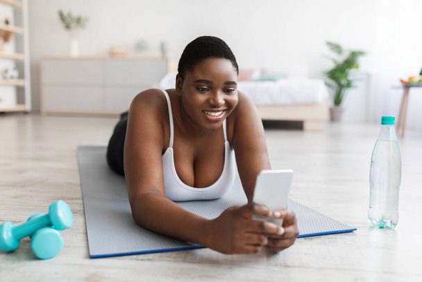 Happy plus size zwarte vrouw met behulp van mobiele telefoon op haar pauze van huishoudelijke training, op zoek naar training video online - Foto, afbeelding