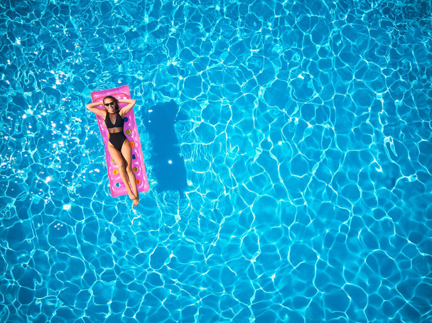 Fit pretty girl in bikini chilling on inflatable pink mattress in swimming pool. Slim hot woman in swimwear tanning. Female relaxing on float in blue water at luxury resort. Aerial, view from above. - Foto, Imagen
