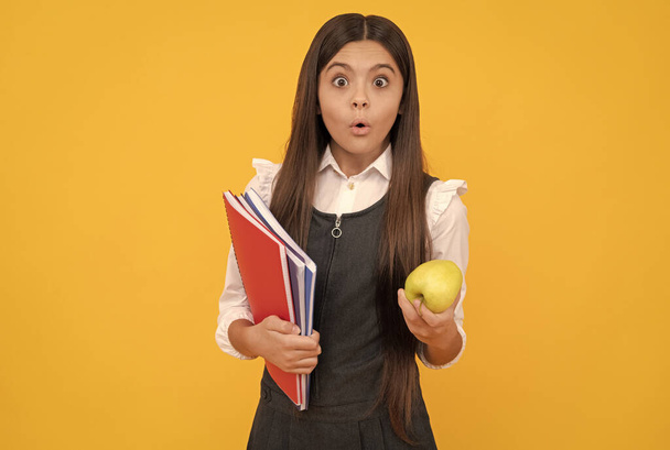 Şaşkın kız çocuğu elma tutuyor ve okul kitapları sarı arka plan, ev ödevi - Fotoğraf, Görsel
