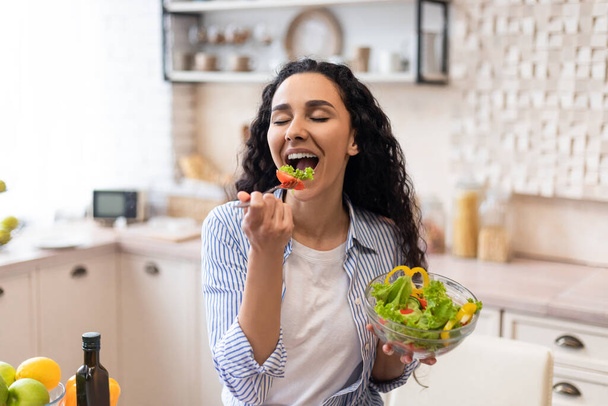 Υγιεινή διατροφική έννοια. Ενθουσιασμένη γυναίκα απολαμβάνοντας φρέσκια σαλάτα λαχανικών, έχοντας δείπνο στην κουζίνα στο σπίτι - Φωτογραφία, εικόνα