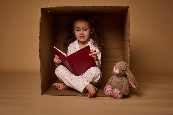 Smiling beautiful child, Caucasian positive pleasant baby girl in pajamas with two ponytails inside a cardboard box, looking at camera isolated over beige background with copy space for ads - 写真・画像