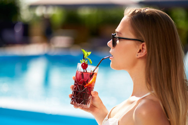 Blonde woman drinks refreshing cocktail sunbathing and sitting near swimming pool at tropical spa. Female in sunglasses enjoys a drink on poolside. Girl chilling in tropical resort on vacation. - Fotoğraf, Görsel
