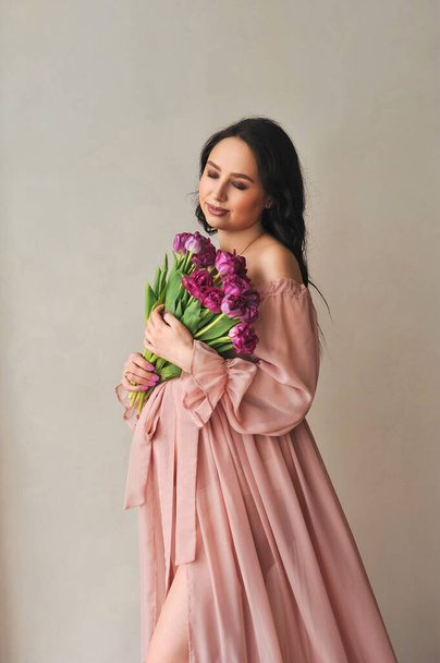 Retrato de estudio de una hermosa mujer embarazada en vestido rosa sosteniendo un ramo de tulipanes violetas en sus manos y cerrando los ojos. Fotosesión de embarazo - Foto, Imagen
