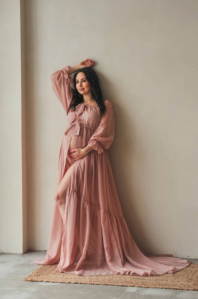 Portret atrakcyjnej kobiety w ciąży w pięknej różowej sukience stojącej przy ścianie w fotostudio i dotykającej ręką brzucha ciężarnej. Koncepcja szczęśliwego macierzyństwa i rodzicielstwa - Zdjęcie, obraz