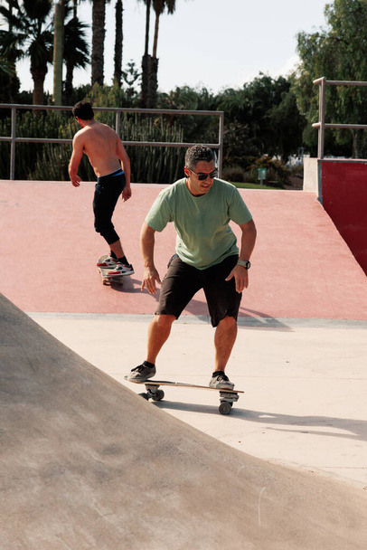 Мужчины катаются на скейтборде в городском скейтпарке. Обычный парень в шортах и футболке. Досуг, спорт экстрим, городской стиль жизни - Фото, изображение