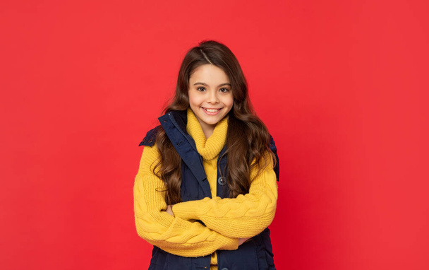 happy child with curly hair in winter down vest on red background, hair care - Foto, Bild