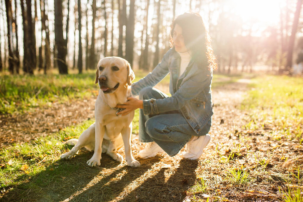 Ein schönes Mädchen umarmt ihren Labrador im Frühlingswald. Junge Frau im Jeansanzug mit Hund bei Sonnenuntergang - Foto, Bild