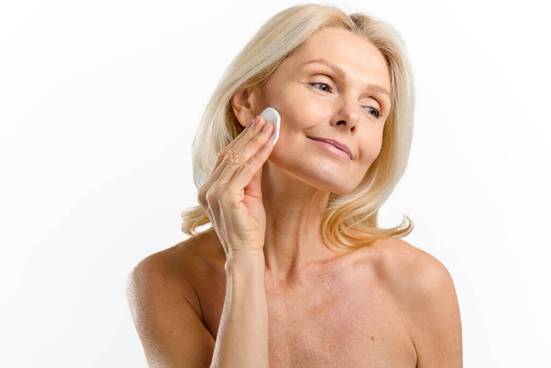 Delighting mid age woman with smooth face skin applying lotion with cotton pad - Fotoğraf, Görsel