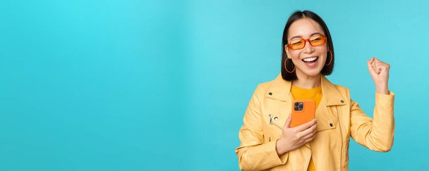 Entusiasta chica asiática sonriente en gafas de sol, sosteniendo el teléfono móvil y bailando, riendo con teléfono inteligente, de pie sobre fondo azul - Foto, Imagen