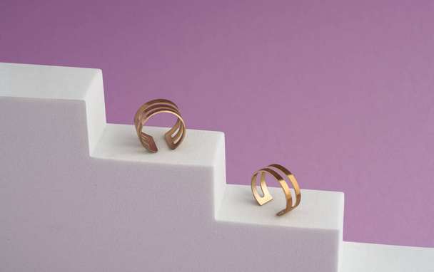 Two modern golden rings on white stairs on pink background with copy space - 写真・画像
