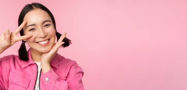 close-up van stijlvolle aziatische meisje glimlacht gelukkig, toont vrede v-teken, kawaii pose, poseren tegen roze achtergrond - Foto, afbeelding