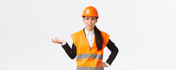 Και λοιπόν, τι έγινε; Απογοητευμένη και απογοητευμένη Ασιάτισσα αρχιμηχανικός παραπονιέται για τους υπαλλήλους, μαλώνει κάποιον, φοράει ανακλαστικό μπουφάν και κράνος ασφαλείας, σηκώνει το χέρι μπερδεμένη - Φωτογραφία, εικόνα