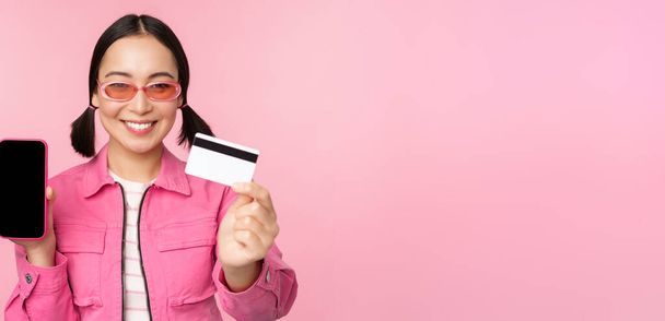 クレジットカードと携帯電話の画面、スマートフォンアプリのインターフェース、オンラインでの支払い、非接触ショッピング、ピンクの背景の上に立つ笑顔の韓国人女性のイメージ - 写真・画像