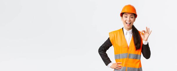 Tyytyväinen onnellinen aasialainen naisinsinööri, rakennuspäällikkö turvakypärässä ja heijastavassa takissa, joka osoittaa hyvää elettä, takaa rakennuksen laadun, varmistaa kaiken erinomaisen - Valokuva, kuva