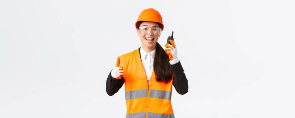 Satisfied happy smiling asian female engineer, industrial technician in safety helmet and uniform showing thumbs-up while praising great work using walkie-talkie, give permission to work - Photo, image