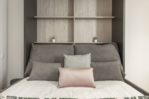 Розкладне ліжко з подушками різних розмірів і кольорів, біла постіль, дерев'яні полиці в студії оренди відпусток
 - Фото, зображення