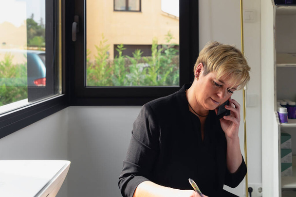 Mature businesswoman working in her home office. Secretary of a beauty salon taking appointments by phone. - Fotografie, Obrázek