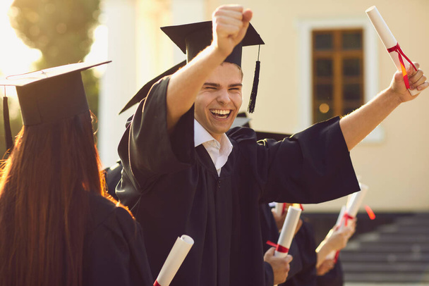 Happy smiling university graduate in mantle holding diploma in raised hand and expressing happiness - Fotoğraf, Görsel