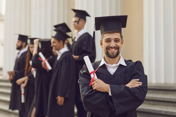 Man in graduation gowns holding diploma and smiling while his friends standing in the background - Fotoğraf, Görsel