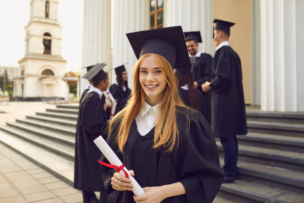 Young smiling girl university graduate in traditional mantle standing and holding diploma in hand - Φωτογραφία, εικόνα