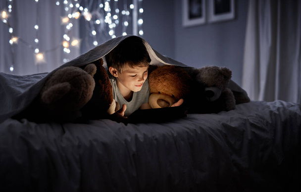 あまりにも眠りに好奇心が強い。夜にベッドでデジタルタブレットを使用して愛らしい男の子のショット. - 写真・画像