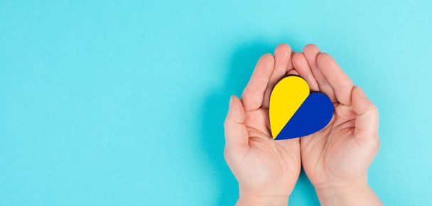 concept of human hands holding heart shaped ukraine flag on blue background. - Фото, изображение