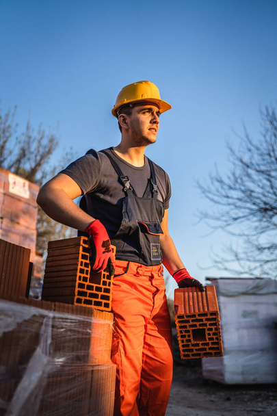 One man construction worker taking and carry or hold clay orange hollow blocks ar warehouse or construction site in sunny summer day copy space - Foto, Imagen