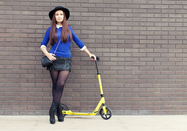 Beautiful brunette girl in a hat with a yellow scooter stands near the brick wall - Fotoğraf, Görsel