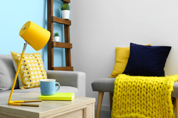 Стол со стильной лампой, чашкой чая и книгой возле дивана в интерьере гостиной - Фото, изображение