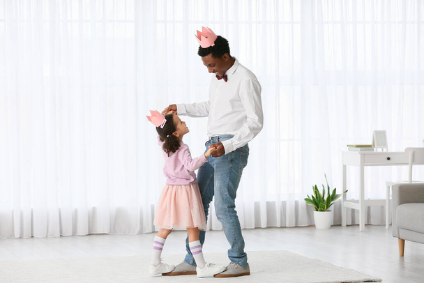 Glücklicher afroamerikanischer Mann und seine kleine Tochter in Papierkronen tanzen zu Hause - Foto, Bild
