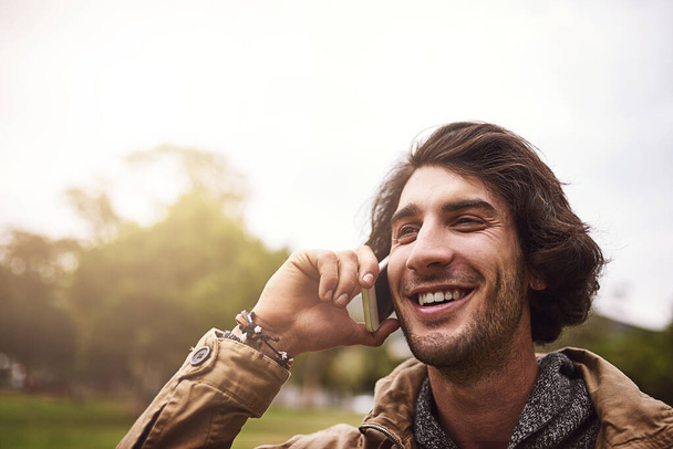 Happy you called. Shot of a cheerful young man talking on his phone while standing outside in a park. - Zdjęcie, obraz