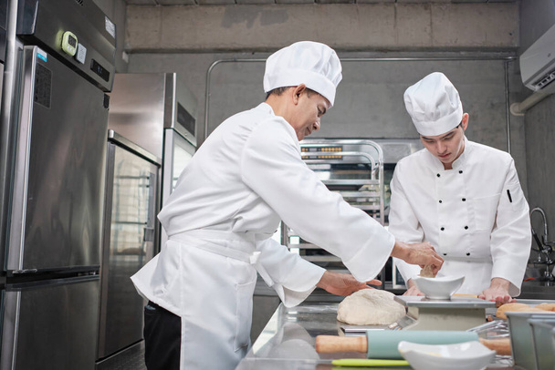 Two professional Asian male chefs in white cook uniforms and aprons are kneading pastry dough and eggs, preparing bread and fresh bakery food, baking in oven at stainless steel kitchen of restaurant. - Foto, Bild