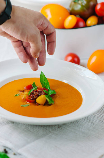 Delicious creamy tomato soup with herbs. Summer light yellow tomato soup. The chef's hand adds a basil leaf to the decor of the dish - Foto, Imagen