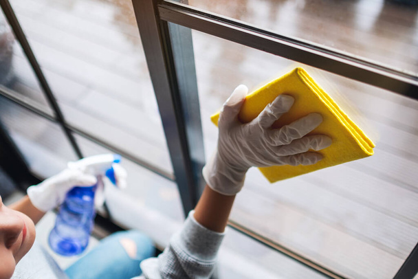 Μια νεαρή γυναίκα που φοράει προστατευτικό γάντι καθαρίζει το παράθυρο για την έννοια της οικιακής εργασίας - Φωτογραφία, εικόνα