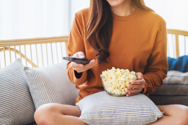 Zamknij obraz kobiety jedzącej popcorn i kanał wyszukiwania z pilotem do oglądania telewizji podczas siedzenia na kanapie w domu - Zdjęcie, obraz