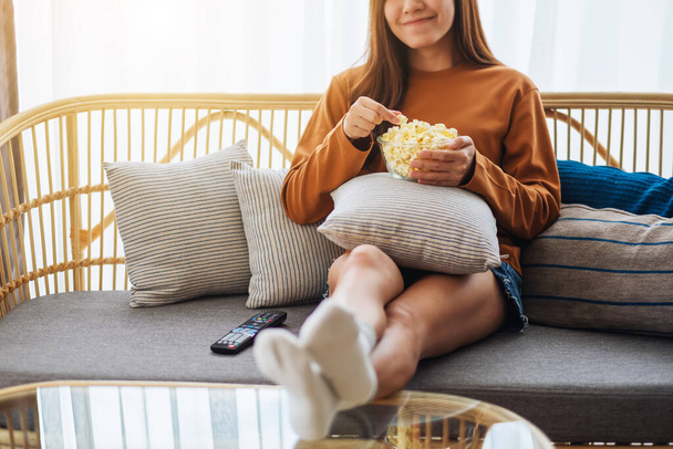 Close up image of a young woman holding and eating pop corn while watching tv and sitting on sofa at home - 写真・画像