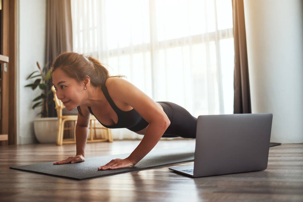 Eine schöne junge Asiatin macht Liegestütze auf der Trainingsmatte, während sie zu Hause Online-Workout-Tutorials auf dem Laptop anschaut - Foto, Bild