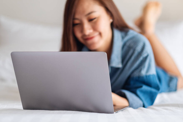 A beautiful asian woman using and working on laptop computer while lying down on a white cozy bed at home - Zdjęcie, obraz