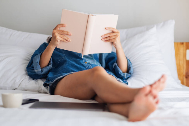 Closeup image of a woman reading book while lying on a white cozy bed at home - 写真・画像