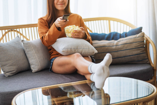 Imagen de cerca de una hermosa mujer joven comiendo palomitas de maíz y canal de búsqueda con control remoto para ver la televisión mientras está sentada en el sofá en casa - Foto, imagen
