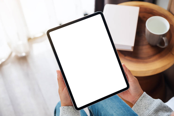 Vista dall'alto immagine mockup di una donna che tiene il tablet pc nero con schermo bianco desktop vuoto mentre si siede su un accogliente letto bianco a casa - Foto, immagini